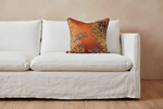 A square decorative throw pillow with an orange and brown patterned design featuring birds and foliage, styled on a white slipcovered sofa against a textured beige wall and wooden floor.