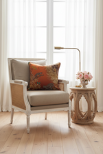 A square decorative throw pillow with an orange and brown patterned design featuring birds and foliage, styled on a white slipcovered sofa against a textured beige wall and wooden floor.