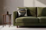 An olive green velvet sofa in a modern living room setting, featuring a square throw pillow with an abstract black, white, and gold design. A small wooden side table with a vase of dried branches sits to the left against a textured gray wall.