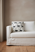 A brown leather armchair with a matching ottoman in a softly lit room, styled with a cream pillow featuring a black cat pattern. A gold floor lamp stands to the left, and a small side table with white flowers is on the right.