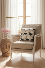 A brown leather armchair with a matching ottoman in a softly lit room, styled with a cream pillow featuring a black cat pattern. A gold floor lamp stands to the left, and a small side table with white flowers is on the right.