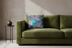 An olive green velvet sofa in a minimalist room setting, styled with a vibrant tropical palm leaf throw pillow and a small side table with a vase of dried branches.