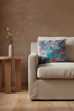 A square throw pillow with a vibrant tropical floral and zebra print in turquoise, pink, and red, styled on a cream-colored sofa next to a wooden side table with a vase of yellow flowers against a textured brown wall.