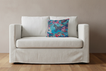 A white slipcovered loveseat sofa with a colorful tropical leaf patterned accent pillow, set against a neutral beige wall and light wood floor.