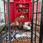 Abstract face-motif rug viewed through French doors in a bold red room with eclectic shelving and a white chair