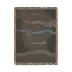 Portrait view of reverse side of abstract woven blanket in dark taupe with teal accent stripe and subtle face, fringe edges