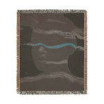Square-cropped back side of abstract woven blanket in dark taupe with lavender-teal stripe and face outline, fringe border