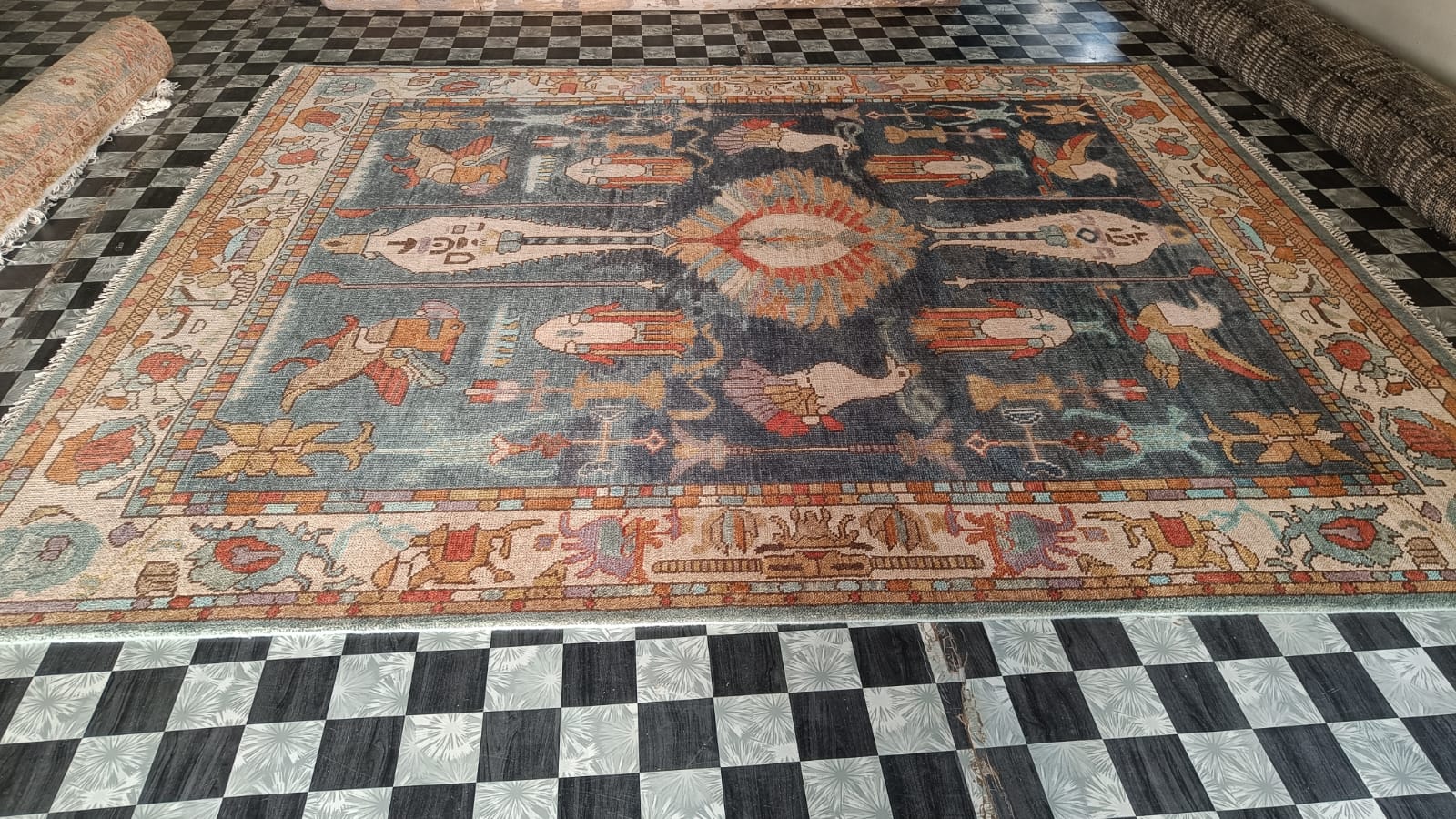 Ancient Wisdom rug laid on a checkerboard showroom floor; dark blue field with Egyptian hieroglyphic figures, birds, cream border, other rugs beside it