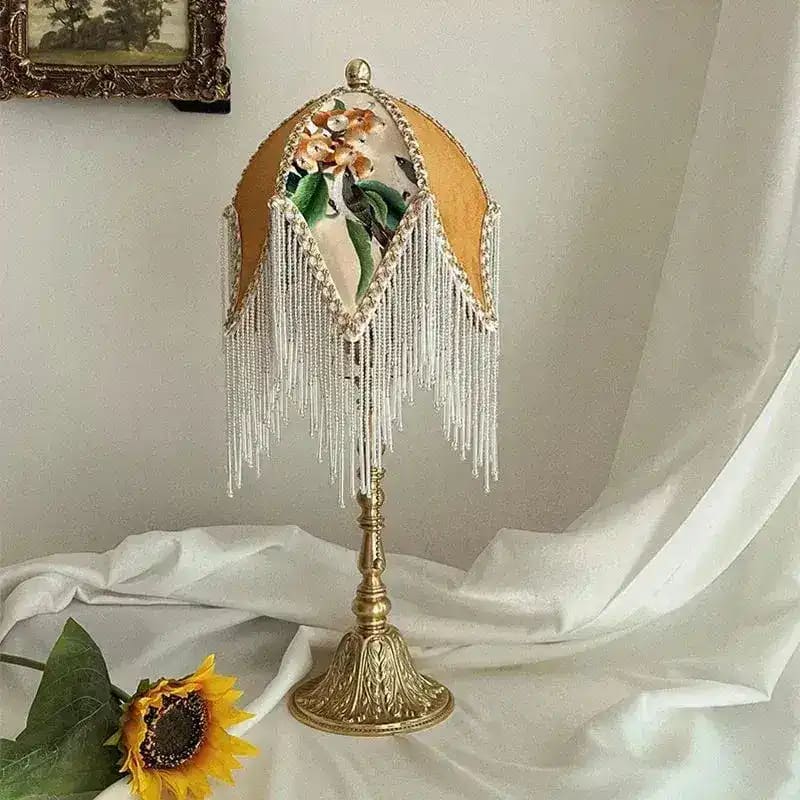Jacquard lamp shade with white beaded fringe on ornate gold table lamp base, white draped background with sunflower