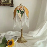 Jacquard lamp shade with white beaded fringe on ornate gold table lamp base, white draped background with sunflower