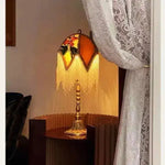 Jacquard lamp shade lit at night — warm amber glow through gold fringe on bedside table, white lace curtain