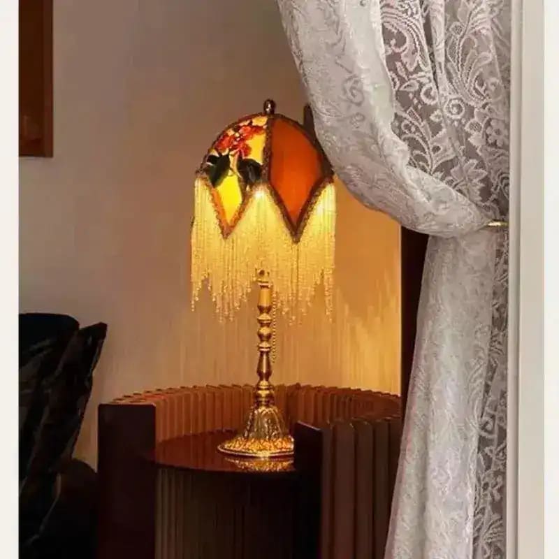Jacquard lamp shade lit at night — warm amber glow through gold fringe on bedside table, white lace curtain