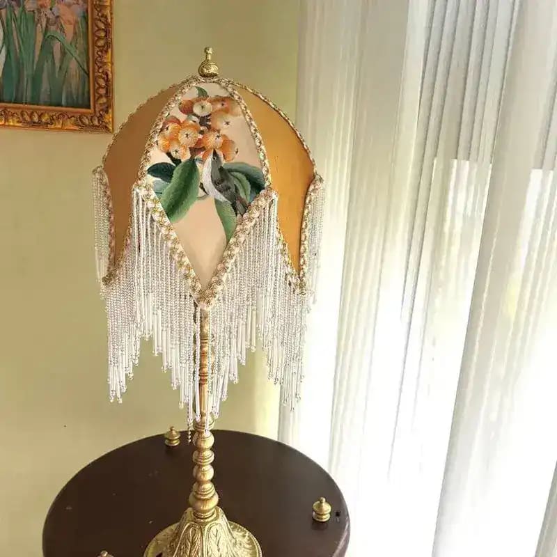 Jacquard lamp shade on gold ornate lamp base on dark side table near window with curtains