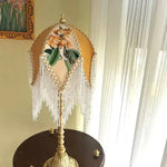 Jacquard lamp shade on gold ornate lamp base on dark side table near window with curtains