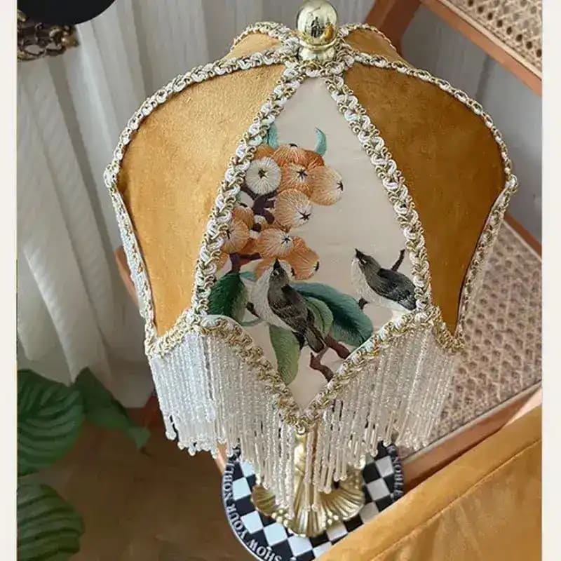 Gold jacquard lamp shade with white beaded fringe, floral bird embroidery panel, in-room setting with rattan chair