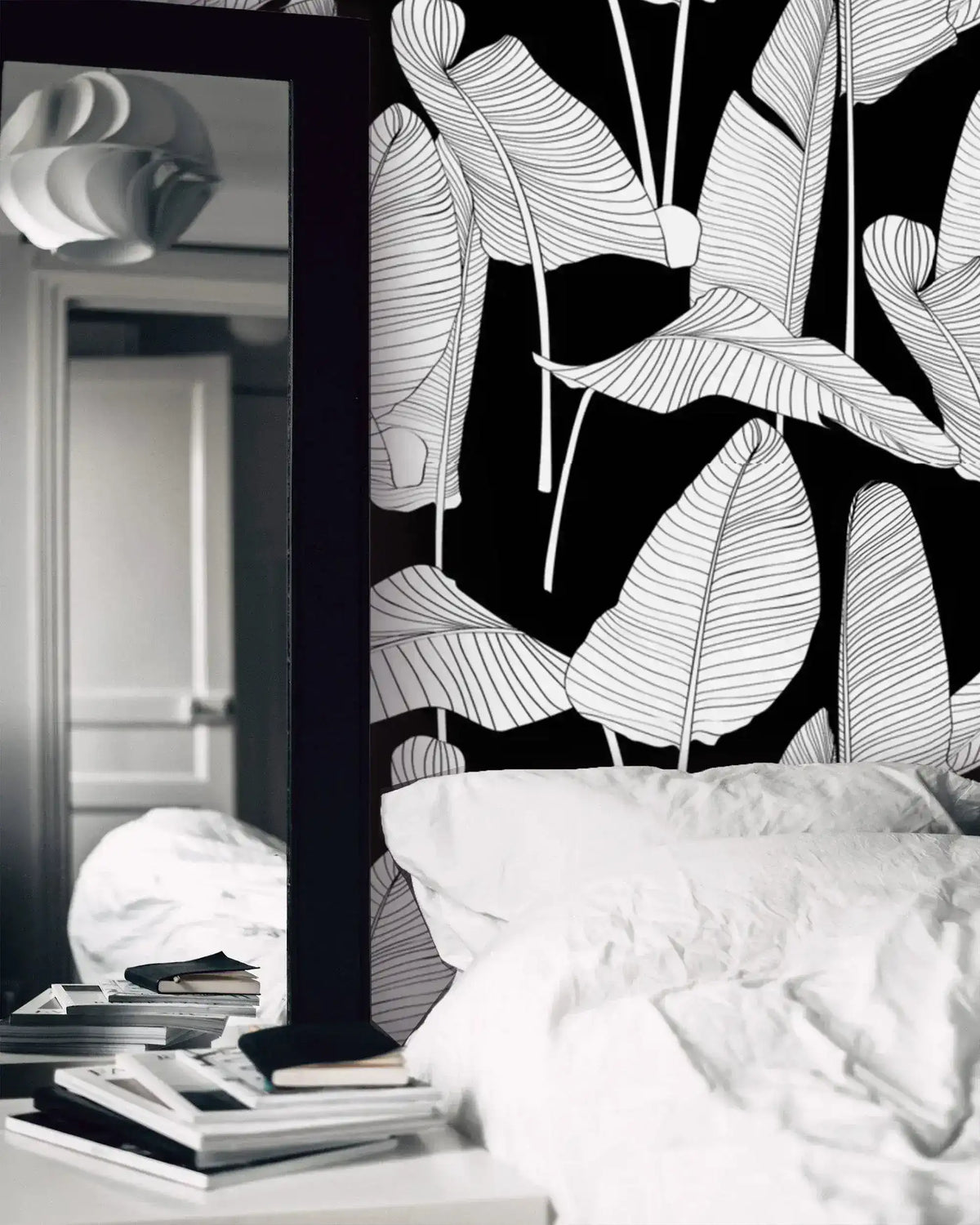 Black and white banana leaf wallpaper in a bedroom setting with white bedding and a mirror nearby