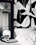 Black and white banana leaf wallpaper in a bedroom setting with white bedding and a mirror nearby