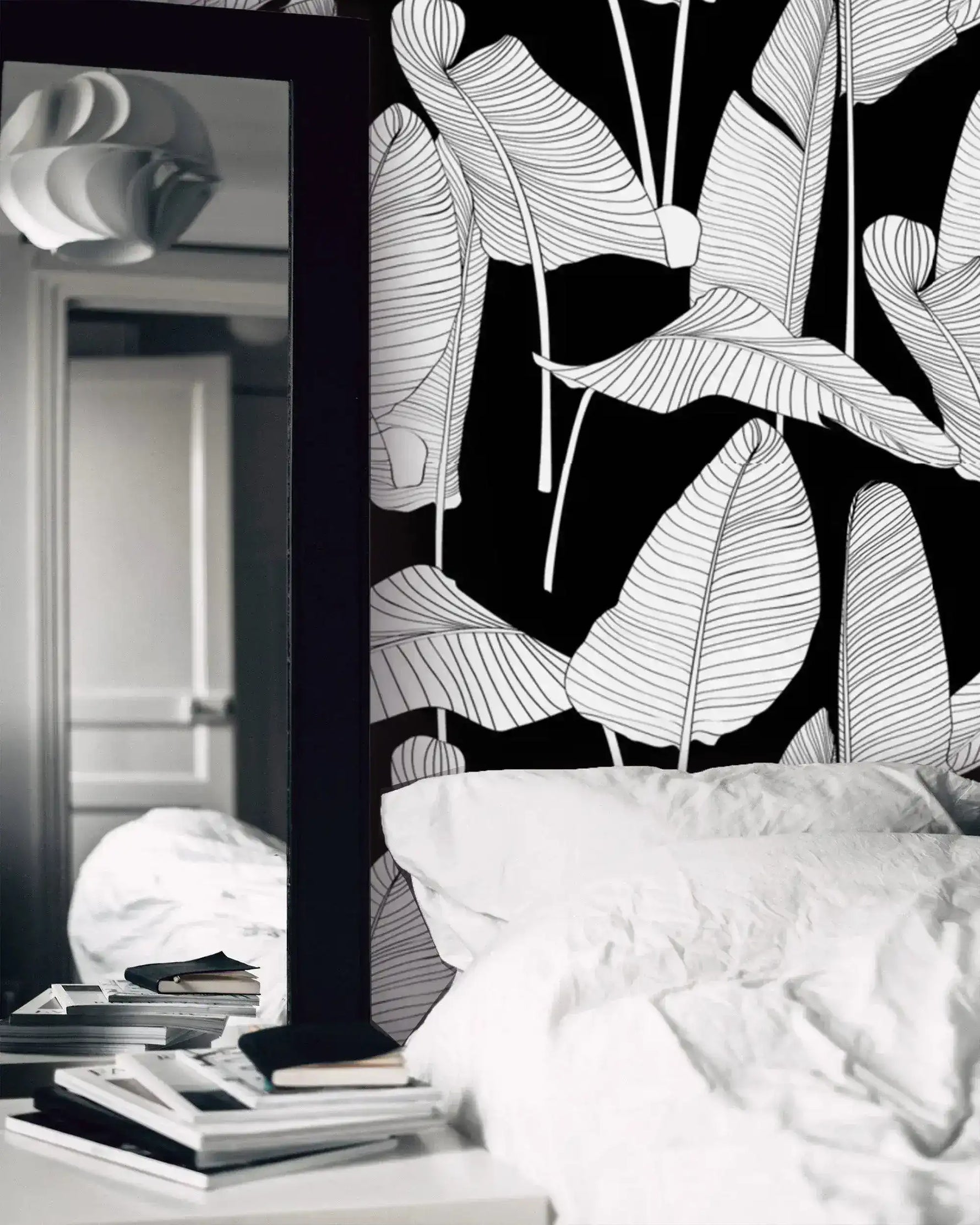 Black and white banana leaf wallpaper in a bedroom setting with white bedding and a mirror nearby