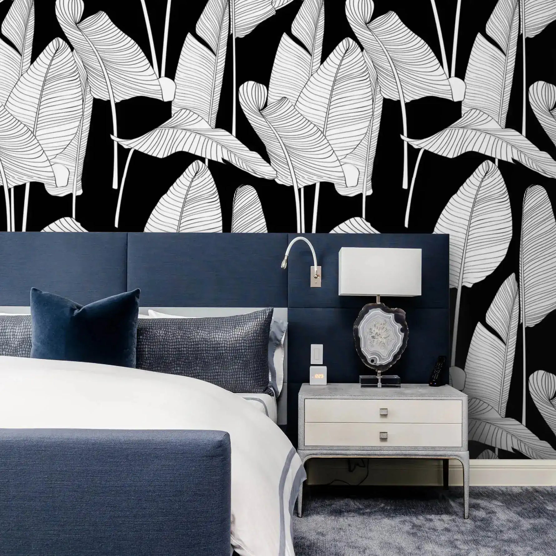 Black and white banana leaf wallpaper above a navy upholstered bed with white bedding and silver nightstand