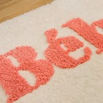 Close-up of a beige bathroom area rug featuring the word 'Bebe' in soft, textured pink letters. The rug appears plush and cozy, ideal for adding a touch of warmth and style to your bathroom decor.