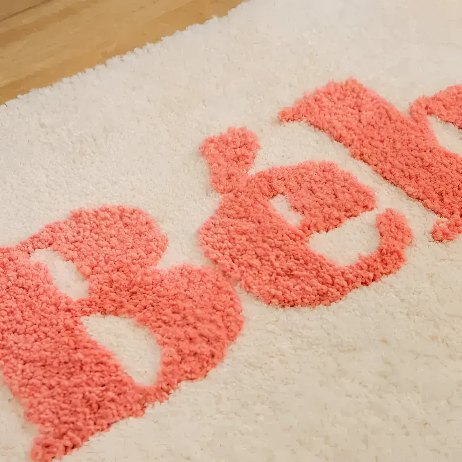 Close-up of a beige bathroom area rug featuring the word 'Bebe' in soft, textured pink letters. The rug appears plush and cozy, ideal for adding a touch of warmth and style to your bathroom decor.