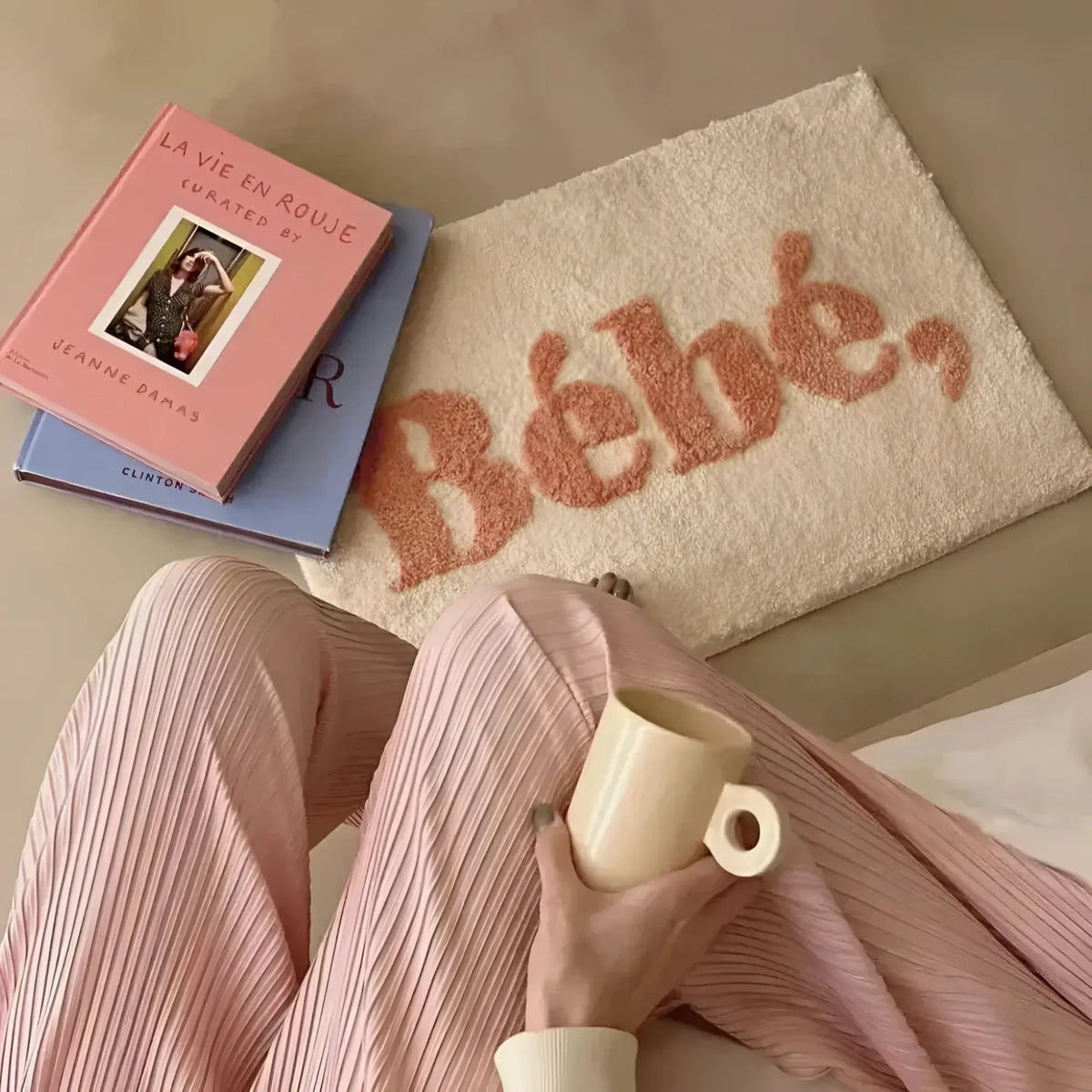A cozy scene featuring an area rug with the word 'Bébé' in pink lettering on a creamy background. The rug is placed on a smooth floor next to a person holding a beige mug, dressed in soft pink pleated pants. Two books, one with a pink cover, are stacked nearby, adding a touch of elegance.