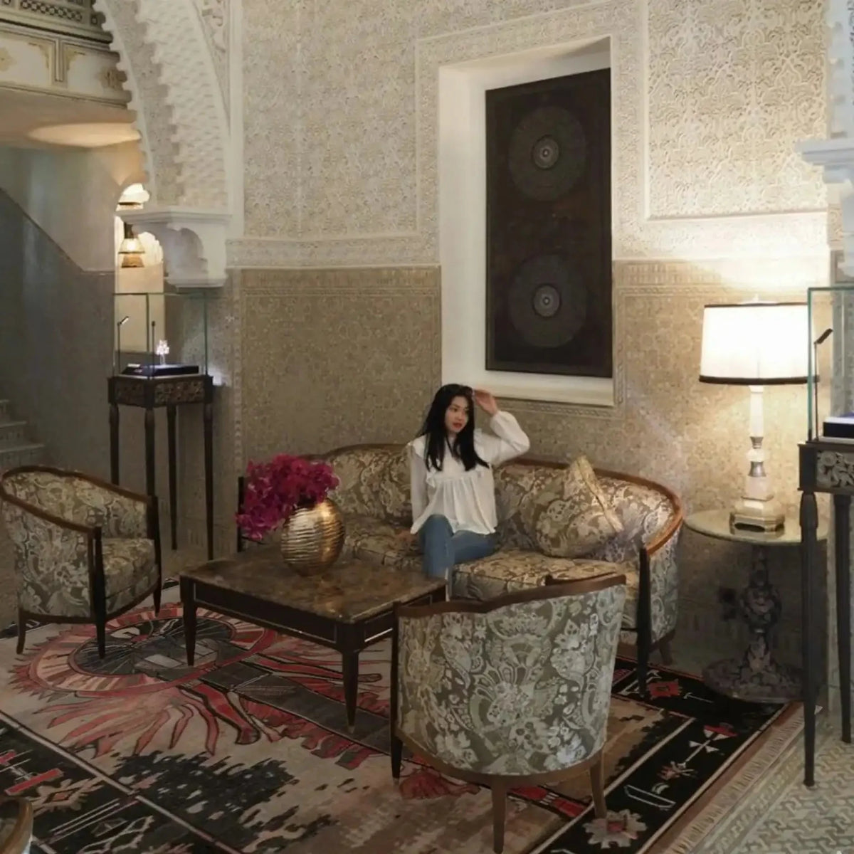 Woman sitting on a couch in an ornate room with intricate wall patterns and decorative furniture.