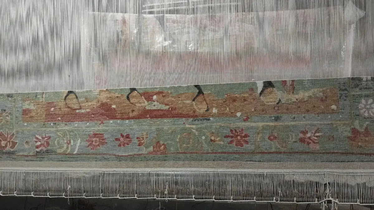 A detailed view of the bespoke hand-knotted wool area rug in progress on the loom, showcasing intricate floral patterns in earthy tones of red, green, and beige.