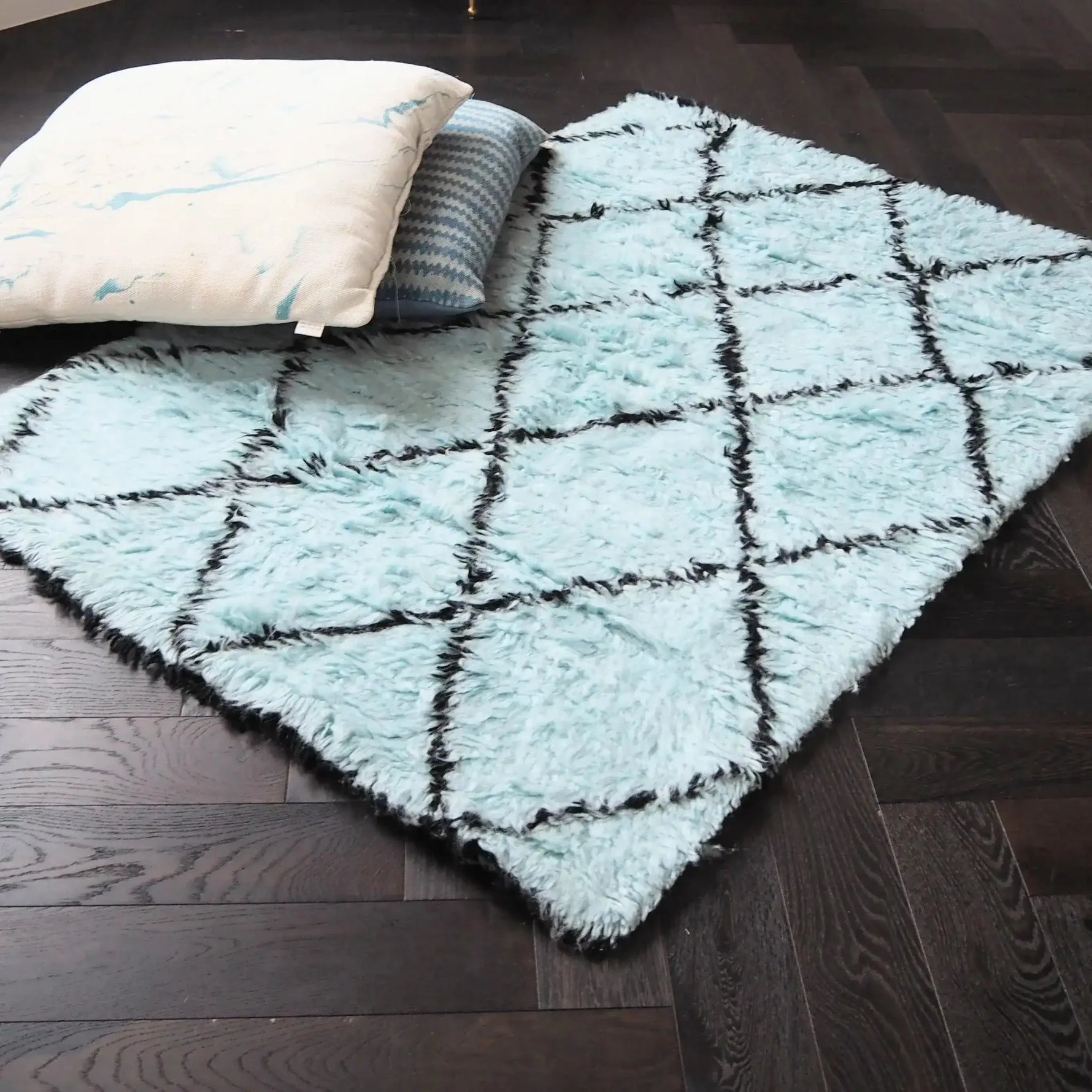 A stylish shag wool area rug featuring a black and blue diamond pattern, adding a touch of elegance to a living room setting. The plush texture of this living room area rug complements the dark wooden floor, while two decorative pillows rest on top, highlighting its cozy appeal. Perfect for those seeking quality rugs for living room spaces.
