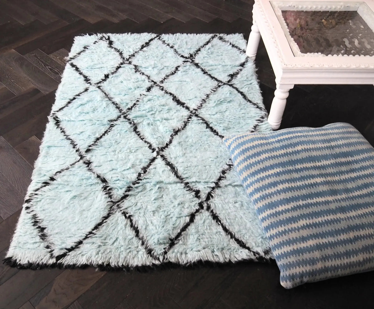 A cozy area rug featuring a black and blue diamond pattern, perfect for enhancing your living room. This shag wool rug adds texture and warmth, complemented by a stylish white side table and a patterned cushion.
