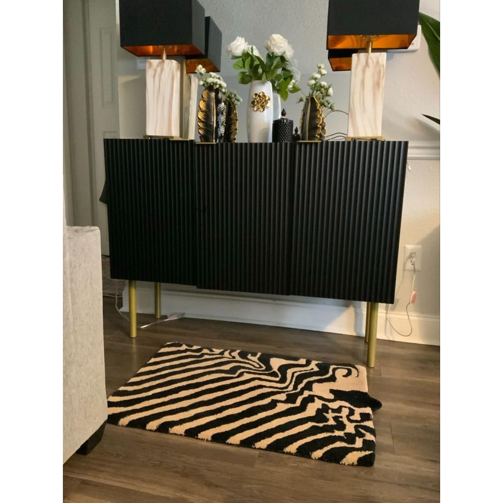 Stylish dining room with a black and tan wool area rug featuring a bold abstract pattern. The rug sits elegantly on wooden flooring, complementing the modern black sideboard adorned with decorative vases and lamps. Perfect for adding a touch of sophistication to any dining table setting.