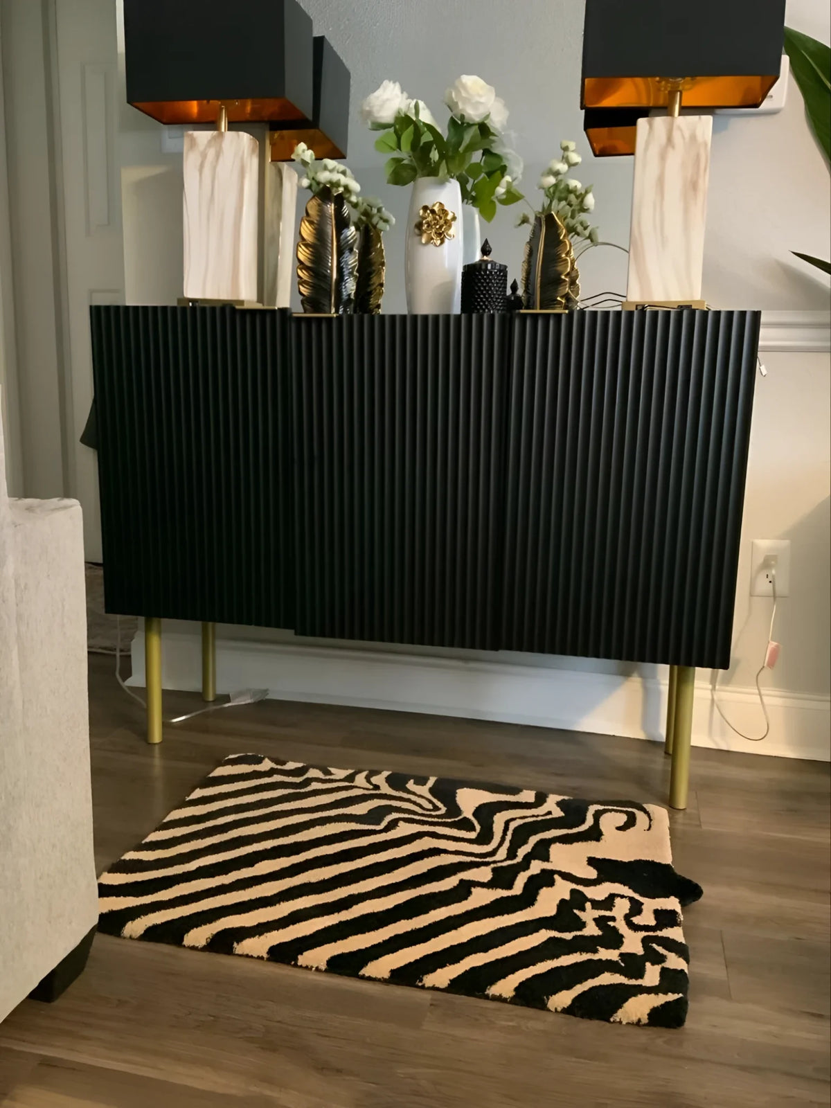 Stylish dining room with a black and tan wool area rug featuring a bold abstract pattern. The rug sits elegantly on wooden flooring, complementing the modern black sideboard adorned with decorative vases and lamps. Perfect for adding a touch of sophistication to any dining table setting.