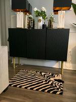 Stylish dining room with a black and tan wool area rug featuring a bold abstract pattern. The rug sits elegantly on wooden flooring, complementing the modern black sideboard adorned with decorative vases and lamps. Perfect for adding a touch of sophistication to any dining table setting.