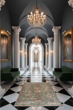 Luxurious hallway with classical architecture, floral rug, chandeliers, and framed portraits.