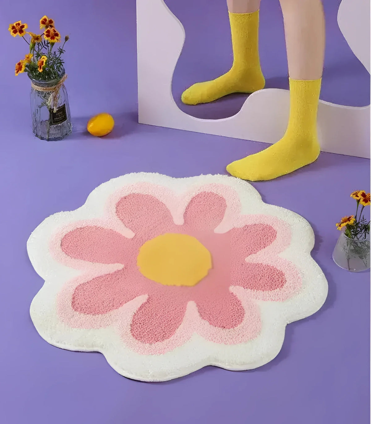 Charming flower-shaped area rug in pastel colors placed on a purple floor. The rug features a pink blossom with a yellow center, adding a playful touch to any room. Yellow socks and vases with flowers further enhance the vibrant setting.