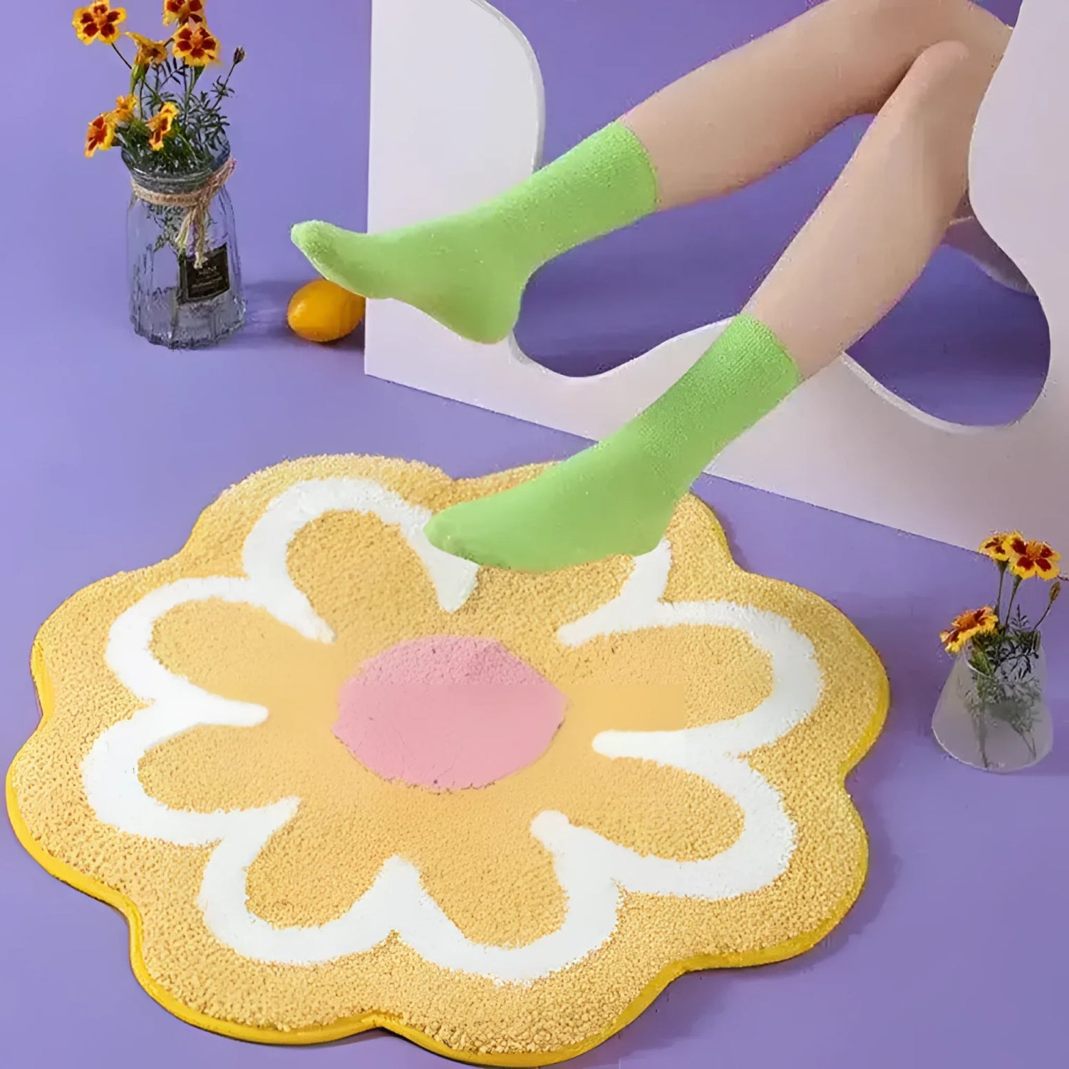 A playful area rug designed like a blossoming flower with yellow petals and a pink center, adding a vibrant touch to any bath space. The rug lies on a purple floor, accompanied by a pair of legs in green socks and small vases with flowers, creating a cheerful and cozy atmosphere.