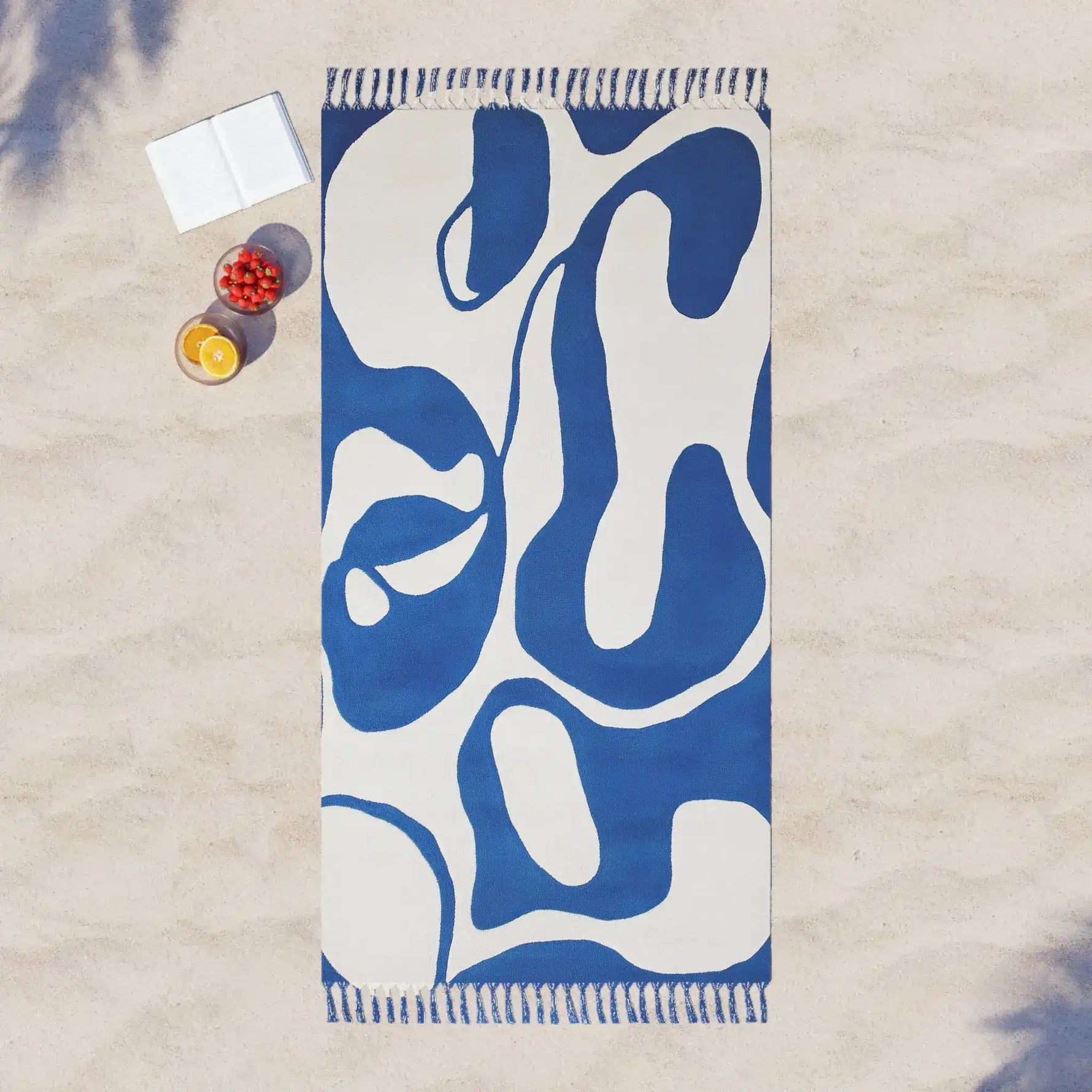 Blue flow abstract beach towel overhead on sand with tasseled ends, white book and fruit bowl props