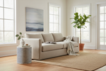 A light beige upholstered sofa in a bright, modern living room with large windows, a woven area rug, a patterned side table, and a potted fiddle-leaf fig plant.