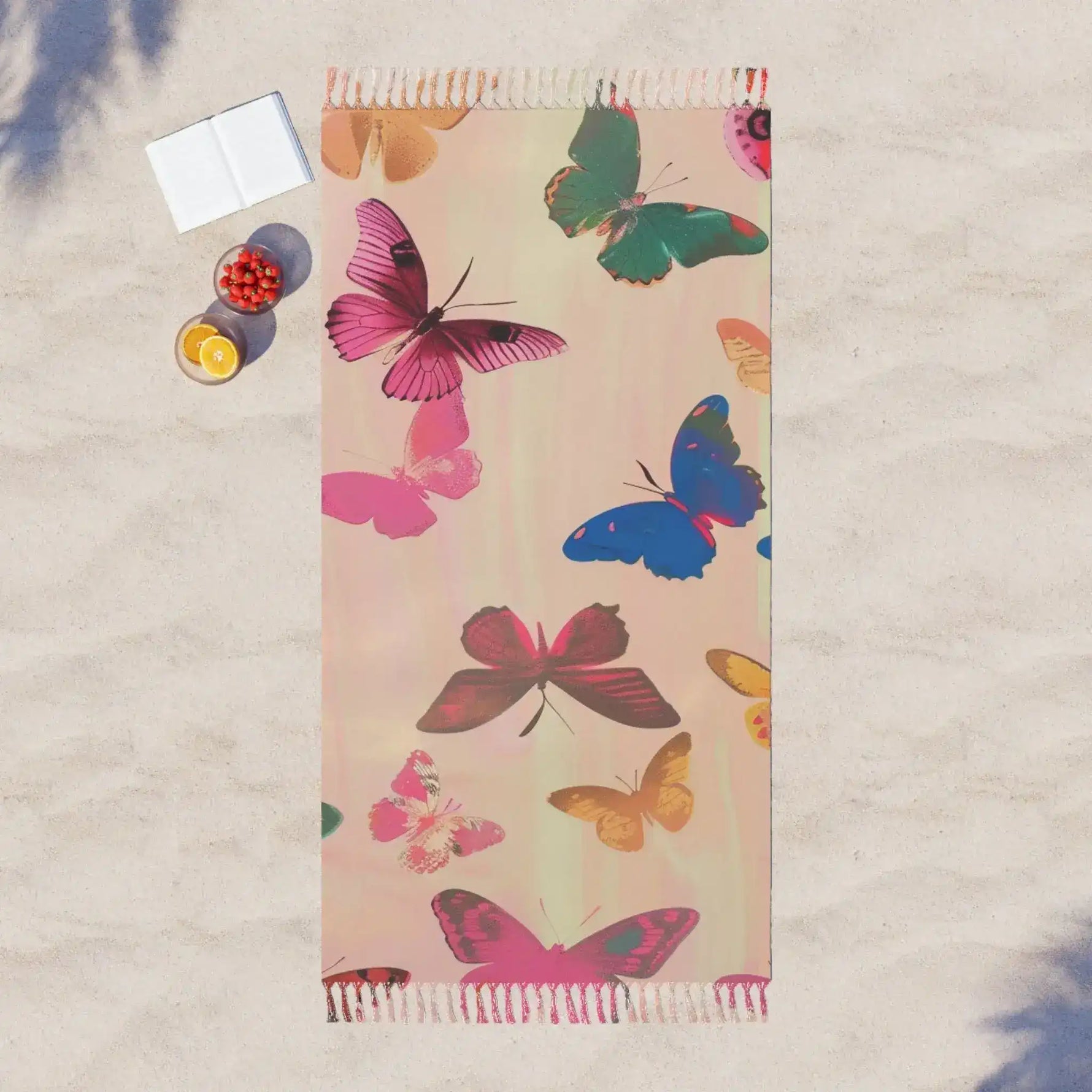 Pink beach towel multicolor butterfly print laid flat on sand aerial view with fruit and book