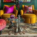 A vibrant living room showcasing a Castle Country Wool Hand Knotted Area Rug, perfect for those seeking vintage charm. The 8x10 area rug features intricate patterns that complement the bold colors of the yellow armchairs and vivid cushions. A unique glass coffee table with artistic metal legs adds a modern touch to the cozy setup, making it ideal for any stylish living space.