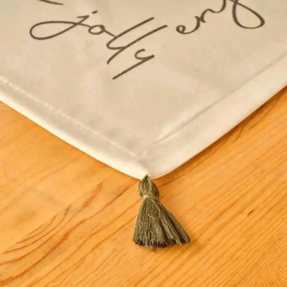 Cheerful Corner Placemats corner detail with olive tassel and festive script text on white cotton against wood surface