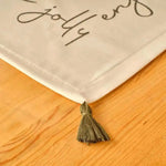 Cheerful Corner Placemats corner detail with olive tassel and festive script text on white cotton against wood surface
