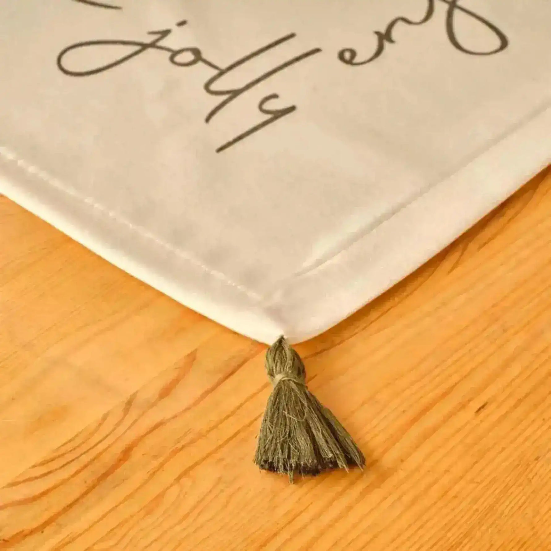 Cheerful Corner Placemats corner detail with olive tassel and festive script text on white cotton against wood surface