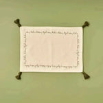 Cheerful Corner Placemats white linen with festive script border and olive corner tassels, flat lay on sage green