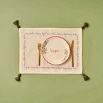 Cheerful Corner Placemats white cotton with festive script border, olive tassels, flat lay with gold cutlery on sage green