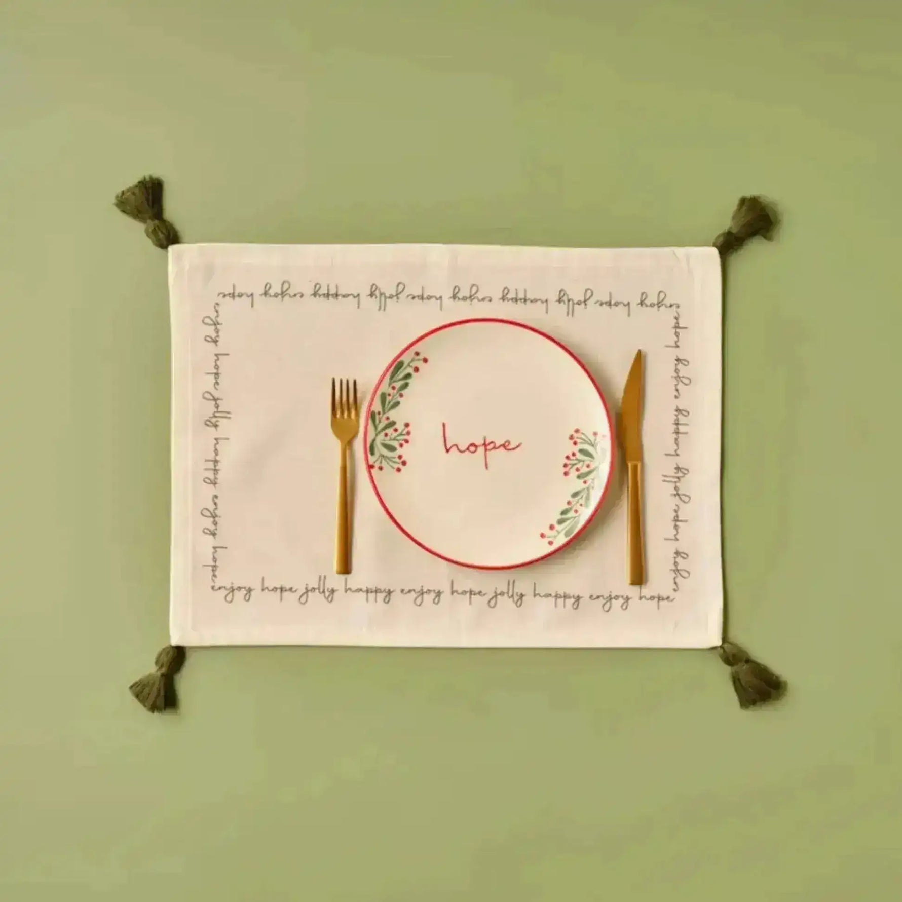 Cheerful Corner Placemats white cotton with festive script border, olive tassels, flat lay with gold cutlery on sage green