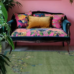 A vibrant living room area featuring a Chinoiserie Peacock in Green Field Wool Hand Knotted Area Rug. The rug lies beneath a colorful vintage sofa with floral patterns and a golden cushion. Lush green plants add a fresh ambiance, making this an ideal setting for rugs for living room decor.