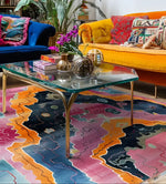 Vibrantly colored area rug with an abstract prismatic pattern, perfect for a contemporary living room. This hand-knotted wool rug features bold shades of pink, orange, and blue, adding a modern touch beneath a glass coffee table and colorful sofas.