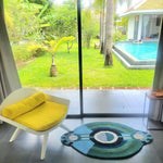 A contemporary round evil eye hand tufted wool rug placed in a modern bedroom setting, featuring a vibrant design with blue and green hues. This stylish area rug adds a pop of color to the space, complemented by a bright yellow chair. Perfect for those seeking unique bedroom area rugs, this large wool rug combines functionality with artistic flair, seamlessly blending with the serene garden view outside.