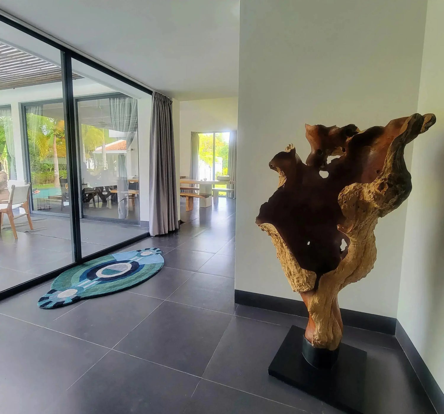 A stylish contemporary round wool area rug with an eye-catching evil eye design sits in a modern living space. The rug adds a touch of elegance and protection, perfect for those seeking unique bedroom area rugs or large area rugs. The room features minimalistic decor with a large window overlooking a garden, enhancing the serene ambiance.